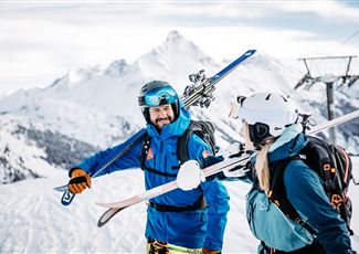 Zwei Skifahrer stehen fröhlich im Schnee und halten ihre Skier in der Hand. Im Hintergrund sind schneebedeckte Berge und ein Skilift zu sehen.