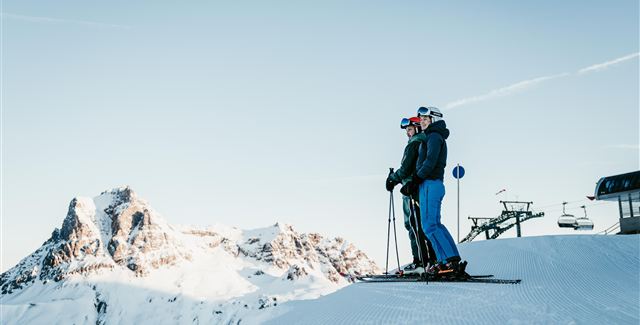 Zwei Skifahrer stehen auf einem verschneiten Hang und genießen die Aussicht auf die Berge. Der Himmel ist klar und blau.