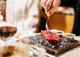 Ein Stück rotes Fleisch wird mit einer Gabel auf einem heißen Stein zubereitet. Im Hintergrund sind Gläser und Saucen zu sehen.