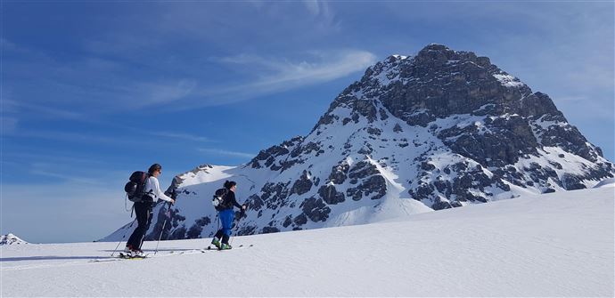 Two skiers on a snowy expanse with a large, rocky mountain in the background. The sky is clear and blue.