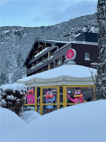 A cozy café with colorful figures in the windows, surrounded by snow. In the background, you can see a modern building and snow-covered mountains.
