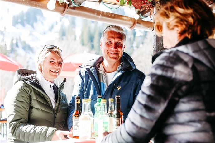 A couple smiles and talks to a waitress at a drinks stand. In the background, mountains and umbrellas can be seen.