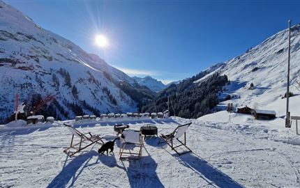 A wintry mountain landscape with snow-covered hills and a bright blue sky. Two deck chairs and a dog are in the foreground.