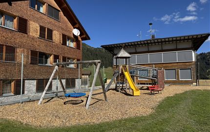 A playground with swings and a climbing tower in a rural setting. In the background, two buildings and blue sky can be seen.
