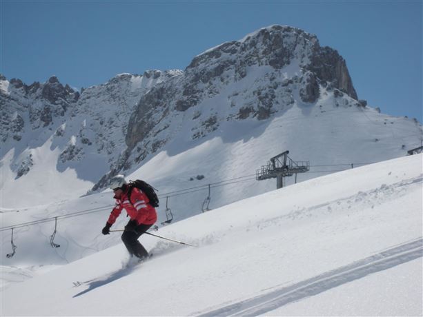 A skier is riding on a snowy slope under a clear blue sky. In the background, there are high mountains and a ski lift.