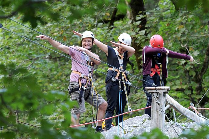 Drie personen in klimuitrusting staan op een touwbrug in het bos en poseren vrolijk. Ze dragen helmen en lijken klaar voor een avontuur.