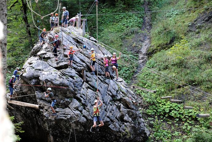 Een groep mensen klimt op een grote rots in de buitenlucht. Omringd door bomen en planten wordt een avontuurlijke buitenervaring weergegeven.