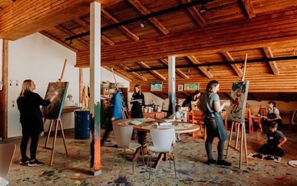 A creative painting workshop with several people working on their canvases. The room has a cozy atmosphere with a wooden ceiling and colorful floor.