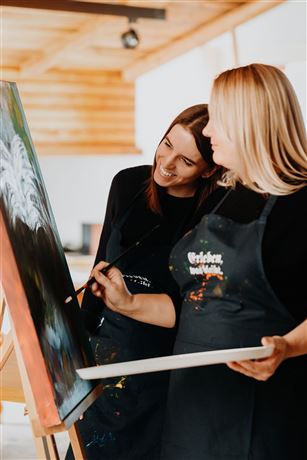 Twee vrouwen werken samen aan een schilderij. Ze dragen schorten en lijken plezier te hebben in het creatieve proces.