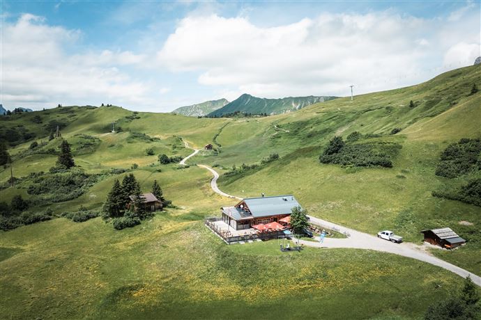 A picturesque mountain lodge surrounded by green meadows and stunning mountains. Two cyclists are riding on the path to the lodge.