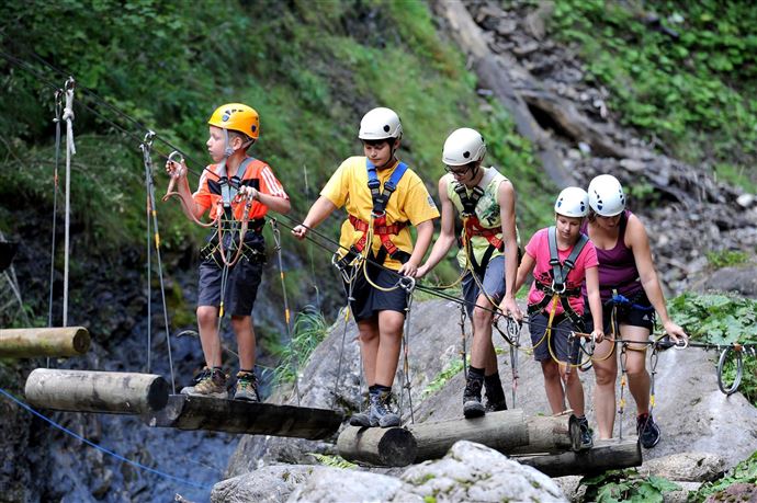 Een groep mensen die aan het klimmen is op een boomstambrug in een natuurlijke omgeving. Iedereen draagt veiligheidshelmen en -harnassen.