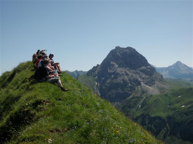 Een groep wandelaars zit op een groene weide met uitzicht op een majestueuze berg. De lucht is helder en zonnig.