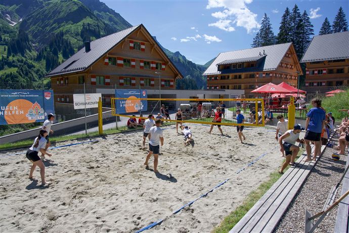 A sand volleyball court with several players in a sunny environment. In the background, there are wooden-clad buildings and green mountains.