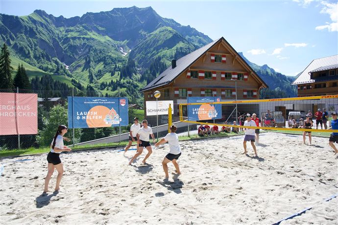 A sand volleyball court in the mountains with several players. In the background, wooden houses and a picturesque landscape can be seen.