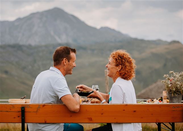 A few enjoying people are sitting at a table outdoors with mountains in the background. They are sharing a meal and smiling at each other.
