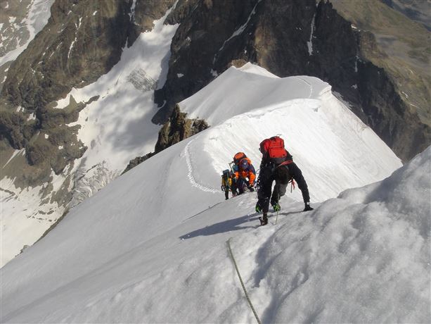 Een groep bergbeklimmers beklimt een met sneeuw bedekte top. Op de achtergrond zijn steile rotsen en berglandschappen zichtbaar.