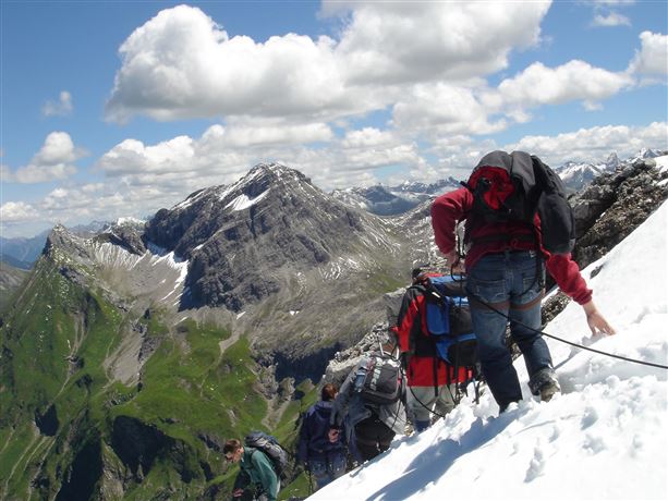 Een groep bergbeklimmers wandelt op een met sneeuw bedekte top. Op de achtergrond zijn indrukwekkende bergen en een bewolkte lucht zichtbaar.