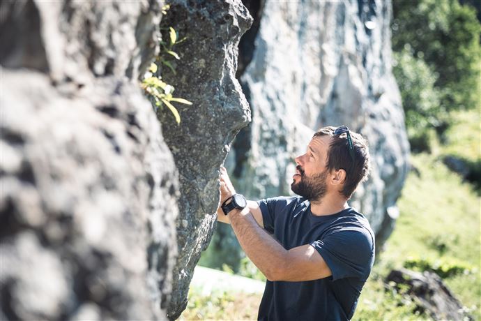 A man is examining a rock in nature. The surroundings are green and sunny, ideal for outdoor activities.