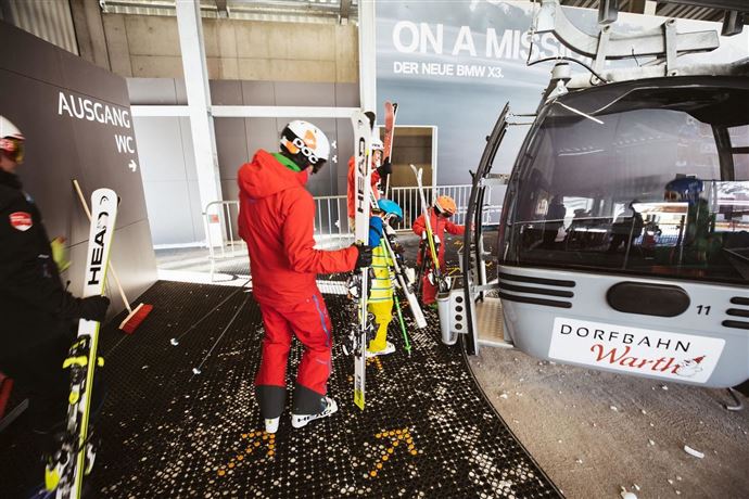 A group of skiers is waiting at the lift to put on their ski equipment. In the background, the cable car leading to the slope can be seen.