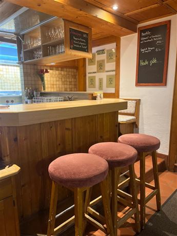 A cozy bar with a wooden counter and three red bar stools. Menus and a chalkboard with special offers hang on the wall.