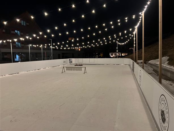Eine beleuchtete Eisbahn bei Nacht mit festlichen Lichtern. Der Platz ist leer und umgeben von Schnee.