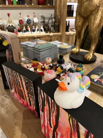 A colorful assortment of rubber ducks and artworks on a table. The central duck has a rainbow mane and stands against a background of decorative pictures.