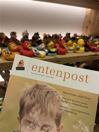 A booklet titled "Entenpost" is lying on a table. In the background, many colorful rubber duck toys are arranged.