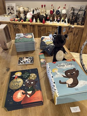 A table with various children's books about "Teddy Eddy" and a stuffed animal in the shape of a bear. In the background, handcrafted decorative items can be seen.
