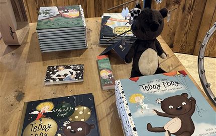 A table with various children's books about "Teddy Eddy" and a stuffed animal in the shape of a bear. In the background, handcrafted decorative items can be seen.