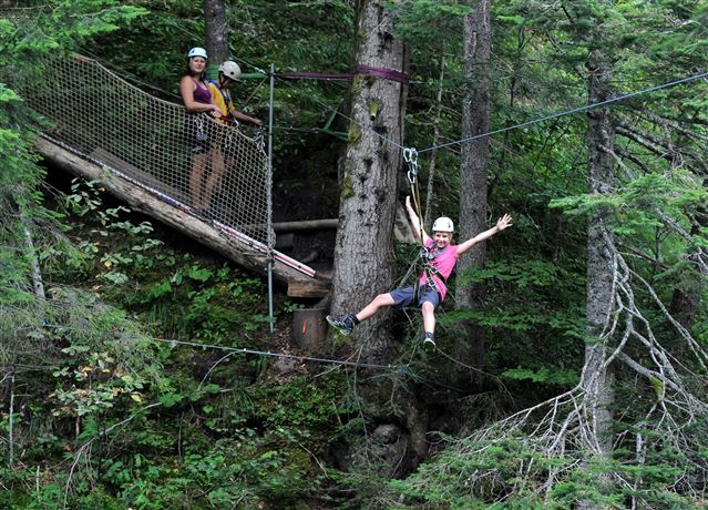 Een jongen zoeft vrolijk langs een zipline, terwijl een vrouw in de buurt toekijkt. Op de achtergrond zijn bomen en een houten platform te zien.