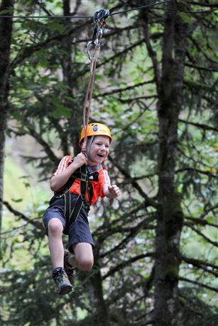 Een jongen gaat met een zipline door het bos. Hij draagt een helm en ziet er opgewonden uit.