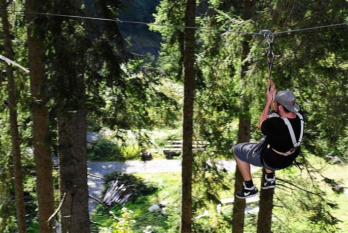 Een persoon zweeft aan een zipline door een bos. Omgeven door hoge bomen en veel groen is de scène helder en uitnodigend.