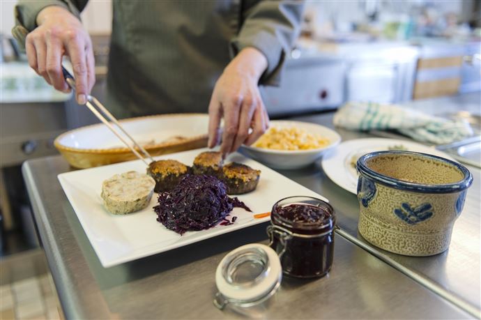 A chef is preparing a dish on a plate that consists of meat and red cabbage. On the table, there are also various side dishes and jars of jam visible.