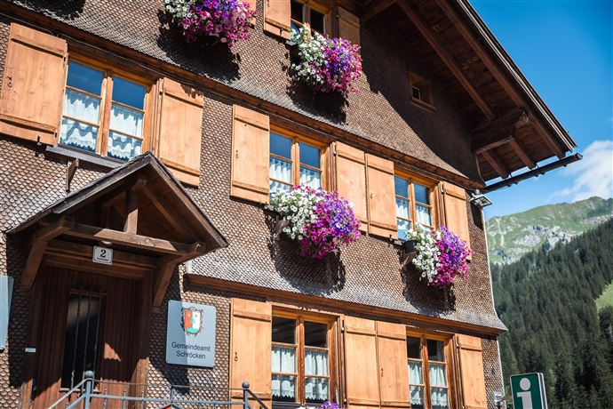 A charming wooden house with colorful flower boxes at the windows. In the background, green mountains and a blue sky can be seen.