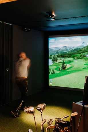 A person is playing golf in a simulator room. In the background, there is a large screen displaying a green golf course landscape.