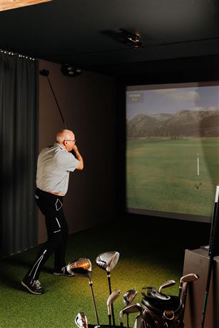 A man is playing golf in an indoor simulator. In the background, a virtual golf course can be seen.