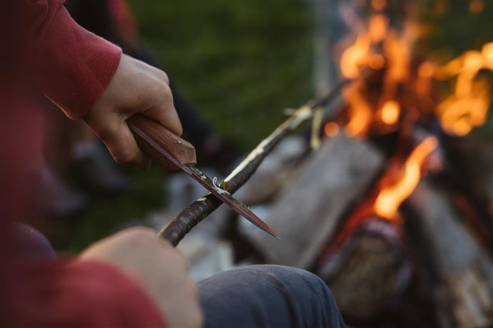 A person is carving with a knife on a wooden stick. In the background, a campfire is burning.