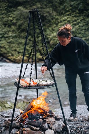 A person is grilling over an open fire by a flowing body of water. The atmosphere is relaxing and close to nature.