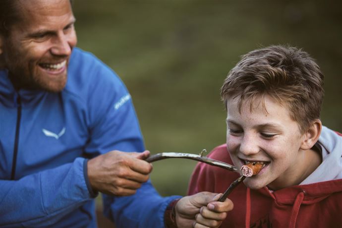 A smiling man is feeding a boy with a piece of food that is attached to a stick. Both are enjoying a friendly moment outdoors.