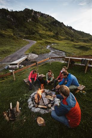 A group of people is sitting around a campfire in nature. In the background, green hills and a path can be seen.