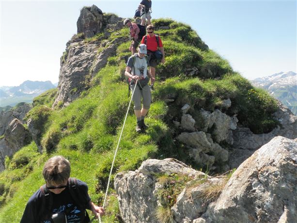 Een groep wandelaars klimt een steile, groene helling op. De omgeving is bergachtig en zonnig, ideaal voor buitenactiviteiten.