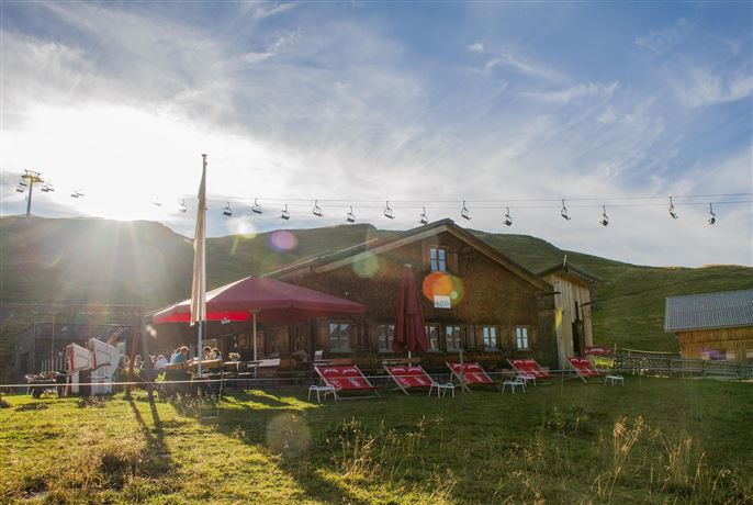 A cozy mountain cabin with a terrace and deck chairs. In the background, there are cable cars and a picturesque landscape.