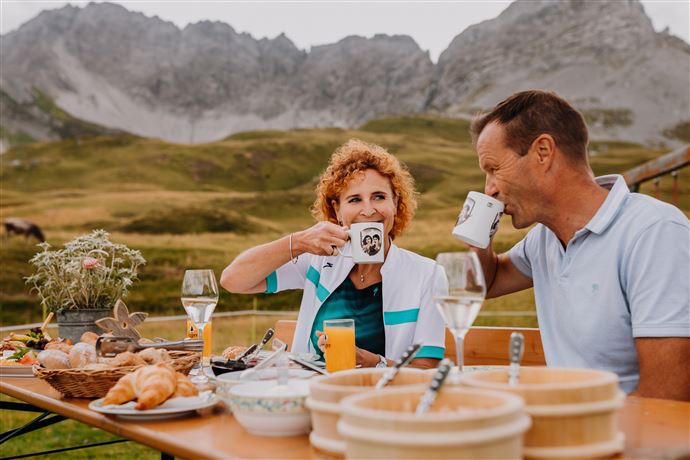 A couple enjoys a shared breakfast outdoors with a magnificent mountain landscape in the background. On the table are drinks, croissants, and flowers.