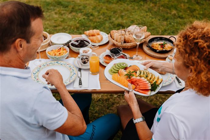 A breakfast table outside with fresh fruits, croissants, and drinks. Two people are sitting across from each other and enjoying their meal in a relaxed atmosphere.