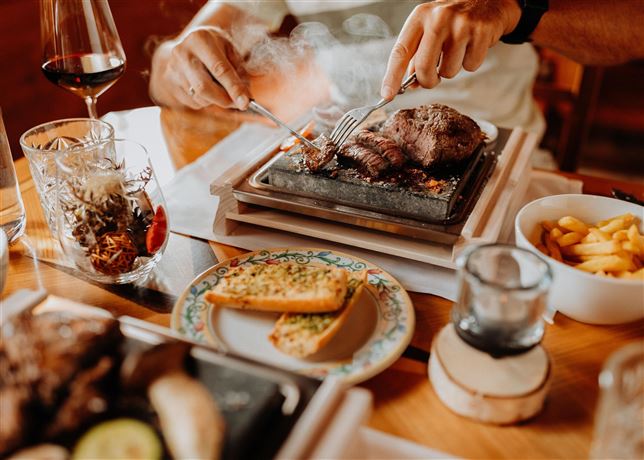 A table with a delicious meal served on a hot stone. There is also a glass of wine and side dishes like bread and potatoes.