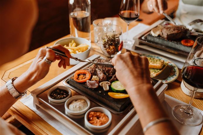 A delicious dish on a hot stone, surrounded by fresh vegetables and various sauces. In the background, drinks and other dishes can be seen.