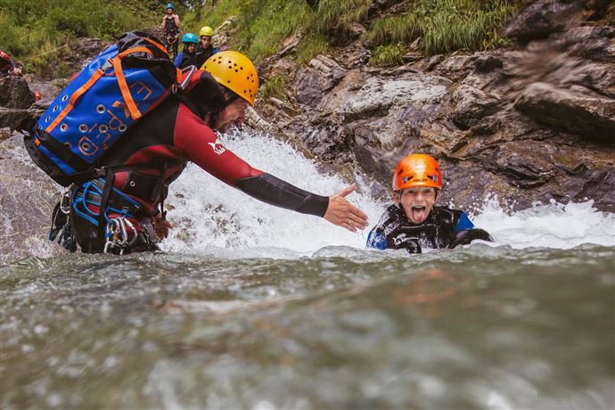 An adventure sports enthusiast is helping another person in the water. Both are wearing helmets and protective gear while being surrounded by rocks.