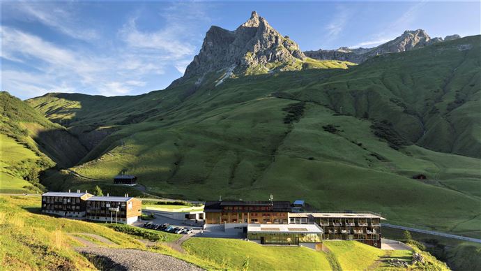 A modern hotel is nestled in a green mountain landscape. In the background, a majestic peak rises under a clear sky.
