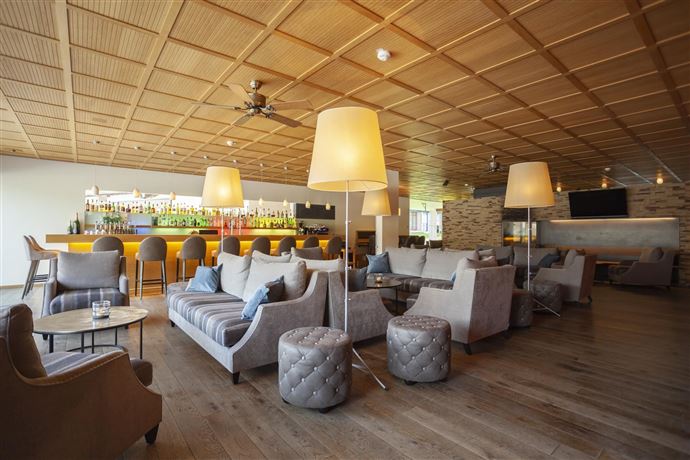 A stylish lounge with comfortable sofas and modern lamps. In the background, there is a bar with various drinks visible.
