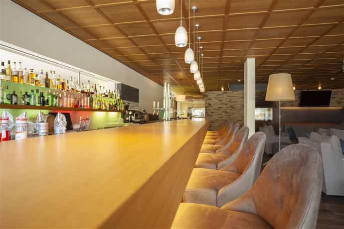 A modern bar with a long wooden counter and elegant chairs. In the background, shelves with various drinks are illuminated.